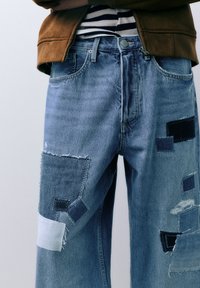 Vervaagde blauwe spijkerbroek met een losse pasvorm, voorzien van verschillende patches in donkerder blauw en wit, en verweerde details op de benen.