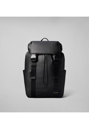 Sort Calvin Klein rygsæk med klaplukning, to forreste stropper med spænder, sidelommer og en logo-tekst på en ensfarvet grå baggrund.