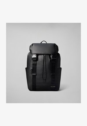 Sort Calvin Klein rygsæk med klaplukning, to forreste stropper med spænder, sidelommer og en logo-tekst på en ensfarvet grå baggrund.