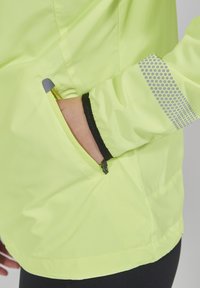 Fluorescenčně žlutá lehká bunda se zipovou kapsou, černým lemováním a reflexními tečkovanými akcenty na rukávu. Hladký povrch.