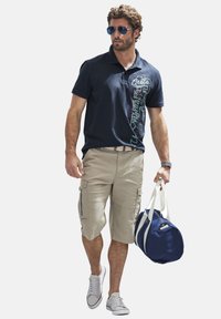 Marineblaues Poloshirt mit grafischem Design, beigefarbene Cargo-Shorts, graue Sneakers und Piloten-Sonnenbrille; hält eine runde, blaue Tasche mit weißen Trägern.