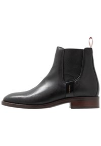 Bottine Chelsea en cuir noir avec panneau latéral élastique, bout arrondi et talon en bois. Dotée d'un logo doré sur le côté et d'une languette à tirer.