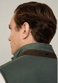 Grüne gesteppte Weste aus strukturiertem Strickstoff, braune Wildlederakzente am Kragen und ein schwarzes Markenlabel auf der Rückseite.