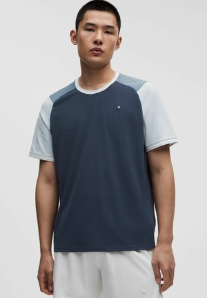 Giovane uomo con maglietta sportiva a maniche corte blu navy e grigio chiaro a blocchi di colore e pantaloncini bianchi, in piedi davanti a uno sfondo semplice.