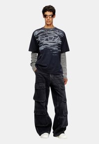 T-shirt graphique bleu marine avec motif de vagues grises et logo, superposé avec des manches longues à rayures grises, associé à un pantalon cargo noir ample et des baskets blanches.