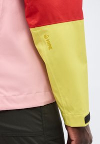 Χέρι ατόμου που φοράει μπουφάν με μπλοκ χρωμάτων με κόκκινα και κίτρινα μανίκια, ροζ κορμό και μαύρο παντελόνι, εμφανίζοντας το λογότυπο "HIPE" στο κίτρινο μανίκι.