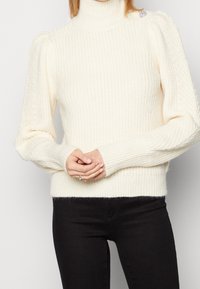 Maglione a collo alto in crema, con maniche a sbuffo e texture a costine, dotato di un girovita aderente e dettaglio decorativo sulla spalla.