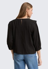 Schwarze Bluse mit Dreiviertelärmeln, lockerer Passform, rückseitiger Schlüsselöffnungs-Detail und strukturiertem Stoff, kombiniert mit hellblauen Denim-Hosen.