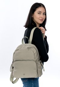 Vaalean oliivinvihreä reppu pehmeästä kankaasta, pyöreällä muotoilulla, vetoketjullisella etutaskulla ja pehmustetuilla olkahihnoilla.