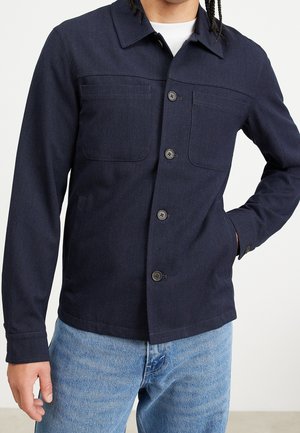 Mann trägt eine dunkelblaue, geknöpfte Jacke mit Brusttaschen und hellblaue Jeans, eine Hand in der Tasche, steht vor einem schlichten Hintergrund.