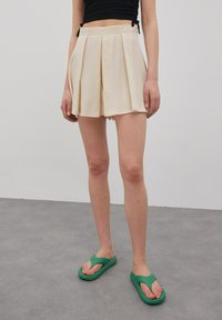 Beige geplooide shorts met een aansluitende taille, gemaakt van lichtgewicht stof, gecombineerd met groene slippers, op een grijze vloer als achtergrond.