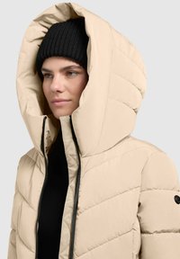 Beige színű pufi dzseki magas gallérral és nagy kapucnival, amely steppelt dizájnnal és fekete cipzárral rendelkezik. Alatta egy bordázott fekete sapka van.