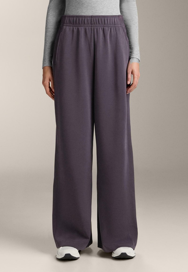 Široké teplákové kalhoty z tmavě fialové tkaniny s elastickým pasem a bočními kapsami, doplněné o bílé sportovní boty.