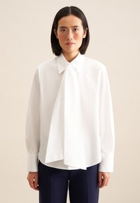Blusa bianca con colletto a cravatta, maniche lunghe e vestibilità rilassata. Tessuto liscio, con orlo leggermente curvato e polsini con bottone.
