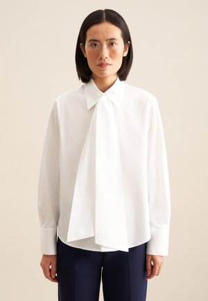 Blouse blanche avec un col à nouer, des manches longues et une coupe décontractée. Tissu lisse, avec un ourlet légèrement arrondi et des poignets à boutons.