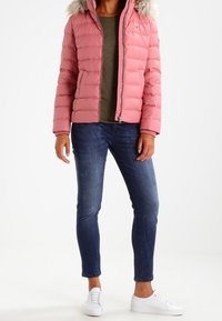 Rosa puffjacka med quiltad design, huva med fuskpäls, dragkedjor, kombinerad med mörkblå smala jeans och vita sneakers.