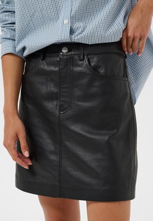 Femme portant une mini-jupe en cuir noire et une chemise bleue à carreaux avec les manches retroussées, les mains reposant près des hanches.