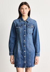 Τζην φόρεμα σε μεσαίο μπλε, με μακριά μανίκια, κουμπιά μπροστά, δύο τσέπες στο στήθος και χαλαρή εφαρμογή με ελαφρώς καμπυλωτό στρίφωμα.