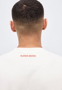 Άνδρας με κοντό, ξεθωριασμένο κούρεμα, που φαίνεται από πίσω, φοράει λευκό πουκάμισο με το "BJÖRN BORG" εκτυπωμένο με κόκκινα γράμματα στο πάνω μέρος της πλάτης.