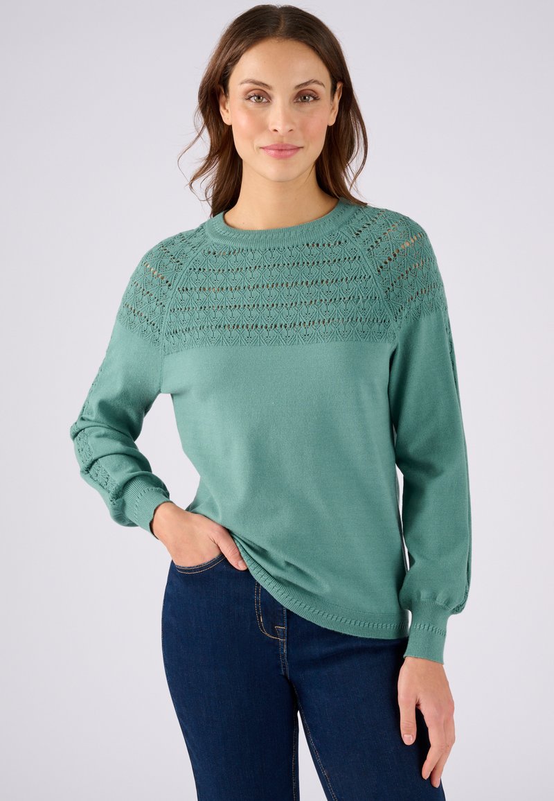 Pull léger vert avec un détail en tricot au niveau des épaules et des manches, col rond et poignets en côtes, porté avec un jean bleu foncé.