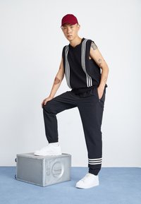 Czarny bezrękawnik w białe paski, czarne zwężane spodnie i białe sneakersy. Model stoi na metalowej skrzyni na jasnym tle.