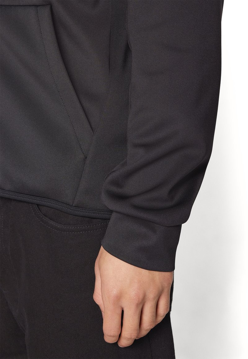 Sudadera deportiva negra con un tejido texturizado, que cuenta con un bolsillo tipo canguro y puños acanalados, combinada con pantalones oscuros.