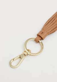 Porte-clé à pampille en cuir marron avec un anneau et un fermoir dorés, présentant une texture plissée et un détail de logo sur l'anneau.