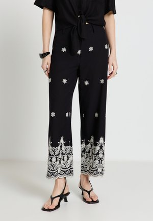Femme portant un pantalon noir à jambes larges avec une broderie florale blanche aux poignets et de petites fleurs dispersées, associé à des sandales noires à talons.