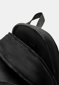 Sac à dos noir avec la fermeture principale partiellement ouverte, montrant un intérieur spacieux et une poignée en tissu sur le dessus.