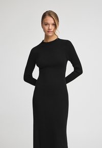 Svart ribbad stickad klänning med långa ärmar och rundad halsringning, med en figursydd silhuett och slät textur.