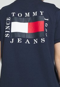 Laivastonsininen t-paita, jossa on näkyvä logo punavalkoisen ja sinisen suorakaiteen muotoisella kuviolla sekä teksti "TOMMY JEANS" ja "SINCE 1985".