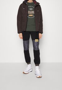 Dark brown puffer jacket, green sweatshirt with logo, black pants with shiny grey panels, and white sneakers with orange accents.