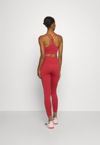 Soutien-gorge sport rosso con spalline incrociate, abbinato a leggings a vita alta coordinati. Il modello indossa sneaker bianche con dettagli arancioni. Texture liscia.