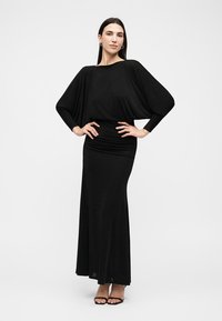 Femme aux longs cheveux foncés portant une robe longue noire à manches chauve-souris et des talons ouverts noirs, debout les mains sur les hanches.