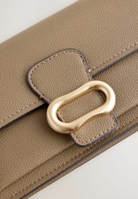 Portefeuille en cuir marron texturé avec une fermeture ovale en or. Les coutures en marron plus foncé soulignent les bords et ajoutent des détails au design.