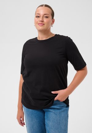 Frau mit heller Haut und zurückgebundenen Haaren trägt ein schwarzes kurzärmeliges Shirt und blaue Jeans, steht mit einer Hand in der Tasche vor weißem Hintergrund.