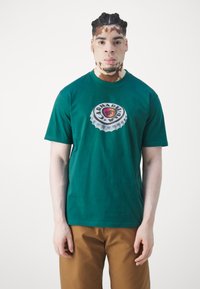 Camiseta de algodón verde con un gran gráfico circular de una tapa de botella con un centro rojo y amarillo y el texto "CARHARTT 100%".