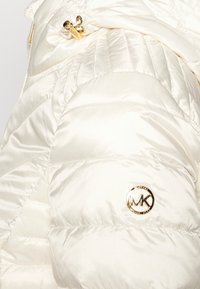 Puchowa biała kurtka z błyszczącym wykończeniem, z diagonalnym przeszyciem, złotym logo oraz złotym zamkiem błyskawicznym.