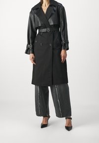 Černý trench coat s koženými rukávy, dvouřadý design, opasek v pase a zvonové manžety. V kombinaci s širokými šedými džíny a špičatými botami.
