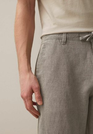 Rechter Arm und Hand einer Person mit beigem Kurzarmhemd und hellgrauer, strukturierter Hose mit Seitentasche und Gürtelschlaufe.