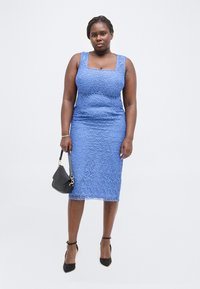 Γυναίκα με μπλε δαντελωτό φόρεμα χωρίς μανίκια κρατώντας μια μαύρη τσάντα, φοράει μαύρα ψηλοτάκουνα με λουράκι στον αστράγαλο και στέκεται μπροστά σε λευκό φόντο.