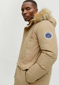 Parka beige con cappuccio foderato in pelliccia sintetica, chiusura con zip, tasche laterali e un patch circolare con logo sulla manica sinistra. Texture liscia.