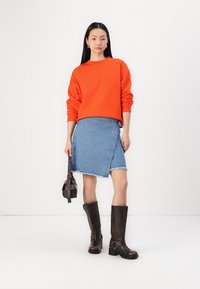 Oransje hettegenser med pregede bokstaver, blå denim omslagskjørt med frayed kant, og brune kne-høye støvler med spenner. Svart crossbody-veske.