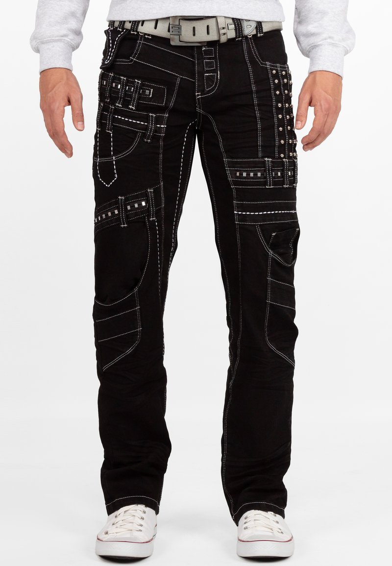 Jeans neri con cuciture bianche a contrasto, numerose tasche e dettagli borchiati. Abbinati a una cintura grigia e sneakers bianche.