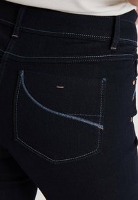 Jeans de mezclilla oscura con costuras azules en contraste, que presentan bolsillos traseros curvados y una textura suave. Se pueden ver sutiles acentos de hardware en tono cobre.