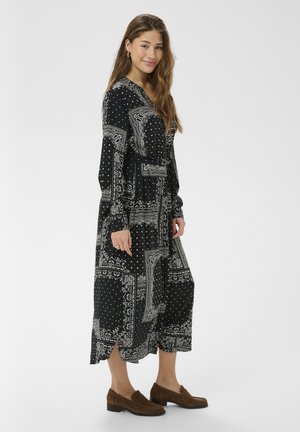 Mladá žena v dlhej čiernej šaty s bielymi paisley vzormi a hnedými mokasínmi, stojaca pred jednoduchým svetlým pozadím.