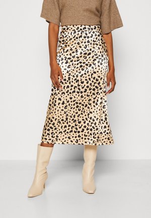 Kobieta ubrana w beżową i czarną midi spódnicę w cętki, brązowy top z krótkim rękawem oraz kremowe botki na obcasie, stojąca na białym tle.