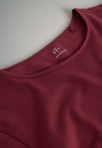 Prim-plan cu un tricou bordo cu guler rotund care arată eticheta interioară cu numele mărcii "The Set" și mărimea medie.