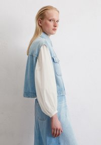 Lichtblauwe denim vest met een hoge kraag, zijzakken en een rafelige zoom, gelaagd over een witte blouse met lange mouwen en geraffineerde manchetten.