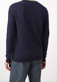 Marinblå stickad tröja med en texturerad mönstring, ribbade ärmslut och nederkant samt rundad halsringning. Buren med ljusgr å byxor.